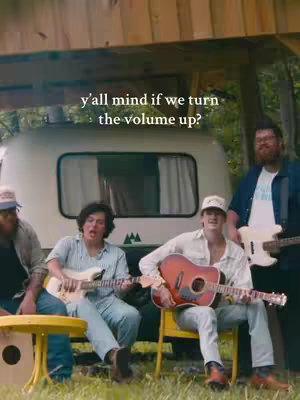 The cover image shows a band of four male musicians positioned in front of a white camper van. The musicians are seated in chairs or on a small stool. They all have guitars and are looking into the camera. The background includes trees and a wooden shelter, giving a sense of an outdoor setting. Text overlayed on the image reads 'y'all mind if we turn the volume up?'