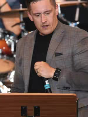 A man in a gray blazer and black shirt stands behind a wooden podium, holding a microphone. The background includes a drum set, implying a live setting.