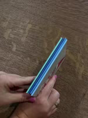 A person's hands hold a stack of Chatbooks, books with various colored edges, resting on a wooden surface. The photo is taken from a slightly high angle, with good lighting. The hands show pink painted finger nails.