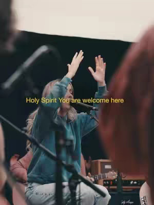 A cover image from a concert shows a person with blonde hair dressed in a turqoise hoodie with their arms raised and their palms facing upward, with the text overlayed.