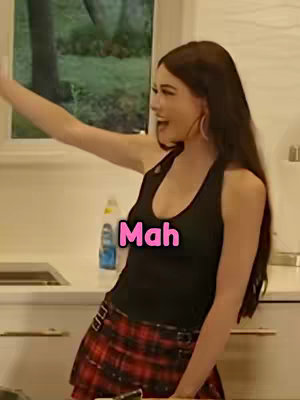 A woman stands in a kitchen, raising her arm and smiling, with a text overlay that reads 'Mah'. She is wearing a black tank top and a plaid skirt. The background is blurry, showing a window and some kitchen elements.