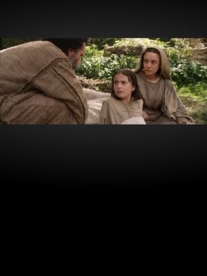 The image shows a scene from a film. Three individuals are seated outdoors. On the left, a man with a beard, dressed in light brown clothing, is partially visible and appears to be speaking to a young boy. In the center, the boy, similarly dressed, looks up attentively. To the right, a woman, also in light brown attire, sits beside the boy with a concerned expression.