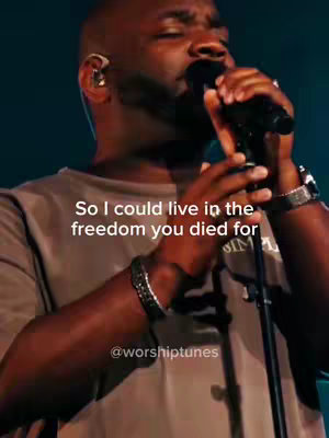 A close-up shot of a Black man singing into a microphone. He is wearing a gray t-shirt and a watch. Text overlayed on the image reads, "So I could live in the freedom you died for". Lighting is focused on the performer. The background is dark.