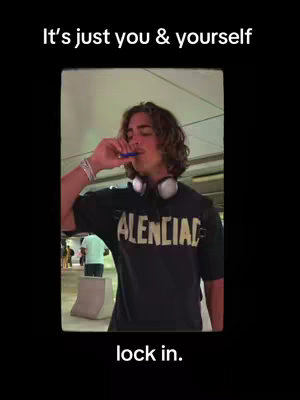 A young man with long, wavy hair is shown in a medium shot, holding a blue object near his mouth as if he's smoking or drinking. He wears a black t-shirt with 'ALENCIAL' in large, yellow block letters. Headphones are around his neck. The background is a blurred interior space, suggesting an airport or public transit area, with lights and a glimpse of other people.