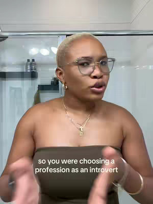 A woman with short blonde hair, glasses, and a strapless top speaks directly to the camera. The background shows a bathroom setting with a mirror and shower visible.