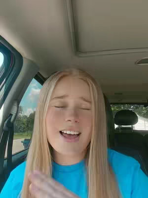 A young woman with long blonde hair is singing in a car, looking up with her eyes closed and her mouth open. She's wearing a light blue t-shirt. The background shows the inside of a car, including the windshield, window area, and the roof.