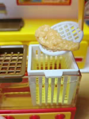 The cover image shows a close-up of a mini fast food kitchen set. A piece of what appears to be fried food rests atop a white basket within the kitchen set. The set's components are predominantly yellow, red, and white. A grid-like tray is visible to the left, with the main focus on the food item and basket.
