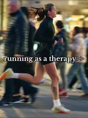 A woman running in a city environment. The background is blurred, suggesting motion, with other people slightly visible. The woman is wearing dark activewear and running shoes, with a ponytail.
