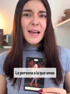 A woman with dark hair and a gray top is centered. She is looking toward the camera with a slightly opened mouth.  She holds a Tarot card, and a white rectangle with black text is laid over it.