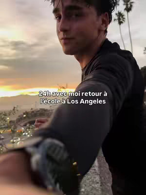 The cover image shows a young man in a selfie-style shot. He is angled slightly away from the camera and looking at it, with a blurred background of a city at sunset. The sky has warm hues of orange and yellows, hinting at dusk or dawn. The foreground features some greenery and a stone or concrete ledge, where the subject is leaning. The man is wearing a dark-colored t-shirt, and the lighting suggests it may be golden hour. Text overlay is present at the bottom.