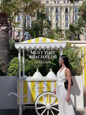 A woman in a white outfit stands by a yellow and white striped cart with a white awning. The background features a large white building and palm trees.