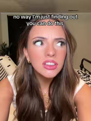 A close-up shot of a woman's face with an expression of surprise or disbelief. She has long, wavy brown hair and is wearing a white top. The background is blurred, with a dark object and a portion of a bed frame visible.