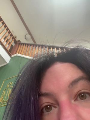 An extreme close-up with the main focus on the eyes of the person. The person has dark hair with strands of purple dye. The background includes parts of a green wall, a staircase, and a white ceiling.