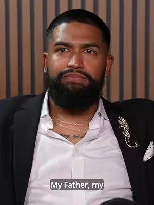 A man of color looking directly at the camera, wearing a suit jacket over a white collared shirt. A necklace and a decorative brooch are visible. He has a groomed black beard.