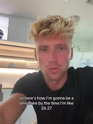 A selfie-style shot featuring a young man with tousled blonde hair. He's positioned center frame and directly facing the camera, giving the impression of a personal video recording. The background consists of various elements: a light-colored wall, horizontal shelves, and hints of what could be a window or another light source creating a space that feels modern and well-lit. Below him is a text overlay describing a goal.