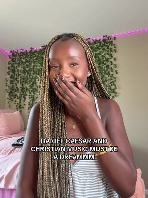 A young Black woman with long blonde braids covers her mouth with her hand, smiling and appearing surprised or delighted. She's wearing a white tank top and gold jewelry. Behind her is a bedroom with a green vine wall decoration and a pink bed.