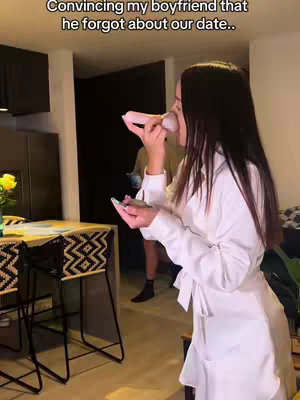 A woman stands in a kitchen, holding a pink object near her face as if speaking into it. Text overlays the image. A man stands blurred in the background.