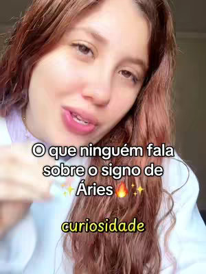 A selfie-style video cover image featuring a woman with auburn hair, looking towards the camera. Text overlays include the question, "O que ninguém fala sobre o signo de Áries" (What nobody says about the sign of Aries), along with the word "curiosidade" (curiosity). The background is a blurred, indoor setting.