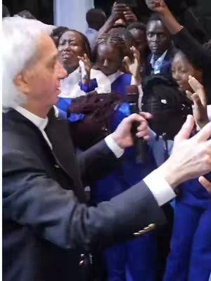 The cover image shows a scene from a religious gathering. A fair-skinned man in a dark suit with a white collar and tie is seen from the side, holding a microphone and addressing a diverse group of people. The background is filled with individuals, including young children and adults, some with their hands raised in a gesture of worship or prayer. The setting appears indoors, possibly a church or event venue. Blue accents are visible on some of the people's clothing. The lighting appears bright and focused, giving a sense of dynamism and energy to the gathering.