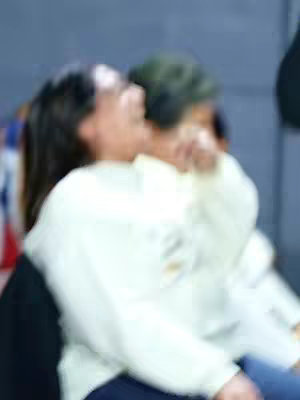 The image has a soft focus, with a person facing left who appears to be in excited state, an other person behind the first one, another person on the right, and blurred colors of blue, gray, and white, suggesting an indoor setting.