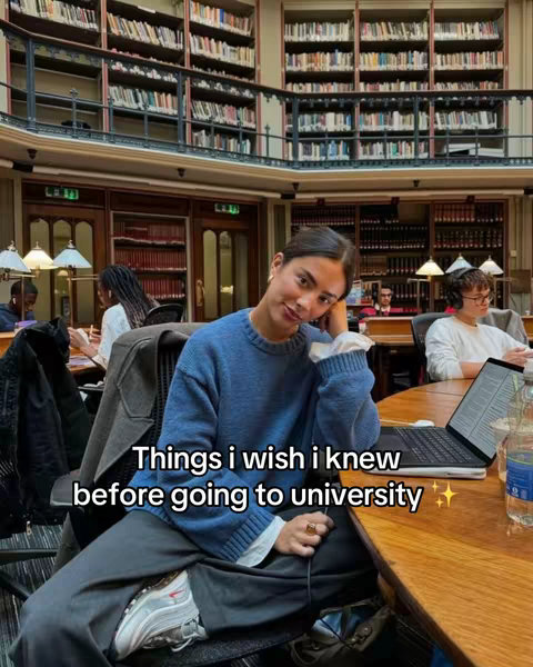 A young woman is seated at a table in a library, writing. Behind her are tall bookshelves filled with books. The woman is wearing a blue sweater and appears to be looking at the camera.