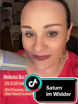 A close-up shot of woman's face with a microphone in the lower left corner, a text overlay at the bottom highlighting "Saturn im Widder". The woman is fair-skinned with dark hair pulled back, adorned with dangling earrings