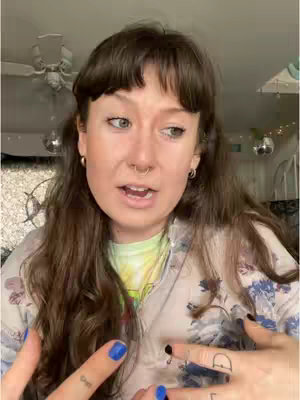 A woman with long, wavy dark hair and bangs is centered in a selfie-style video. She's looking slightly off-camera with a questioning expression, mouth open as if speaking. She is wearing a patterned button-down shirt over a tie-dye t-shirt, with a small gold hoop earring in her ear. She has a nose ring and multiple finger tattoos. Her fingernails are painted blue. The background includes a ceiling fan, decorative orbs, and a glimpse of a window.