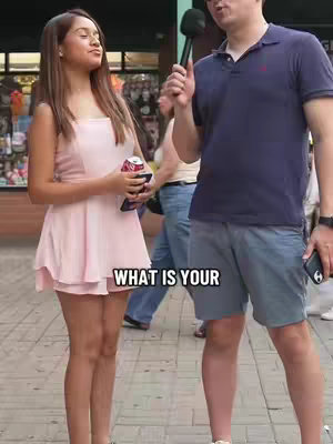 A man holding a microphone is interviewing a fair-skinned woman in an urban environment. The woman, dressed in a light pink dress and holding a can of soda and a phone, looks up and to her left, appearing to listen to the question. A text overlay in black reads "WHAT IS YOUR" in a bold, sans-serif font.