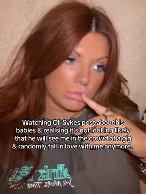 A woman with reddish-brown hair is centered in the frame, her gaze slightly off to the right. The background is a neutral beige. She's touching her lower lip with her index finger, giving a thoughtful or slightly sad expression. Text overlays the picture, informing the viewer about the video's context.