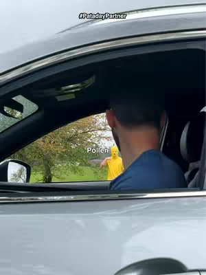 A man in a car looks towards a figure in the environment in a yellow costume that says 'pollen'. The composition is framed from inside the car.