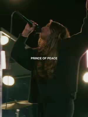 A woman is singing into a microphone on a stage. She has long wavy hair and is wearing a dark sweater. The background is blurred, but appears to be a live performance with stage lights. Text overlayed on the image reads "PRINCE OF PEACE". The lighting creates a warm glow around her.