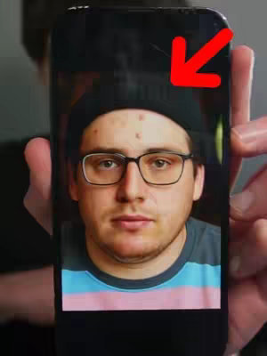 A person is holding a phone displaying another person's face, with a red arrow pointing at the top of the phone. The person on the phone's screen is wearing glasses, a beanie, and a striped shirt. The background is blurred and a hand is visible.
