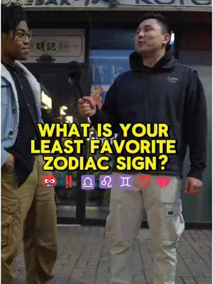 Two men stand on a sidewalk, facing each other. The man on the left wears glasses and a light-colored jacket over a black shirt. The man on the right holds a microphone and wears a black vest. The text overlayed on the image reads, "WHAT IS YOUR LEAST FAVORITE ZODIAC SIGN?" The background suggests an urban setting.