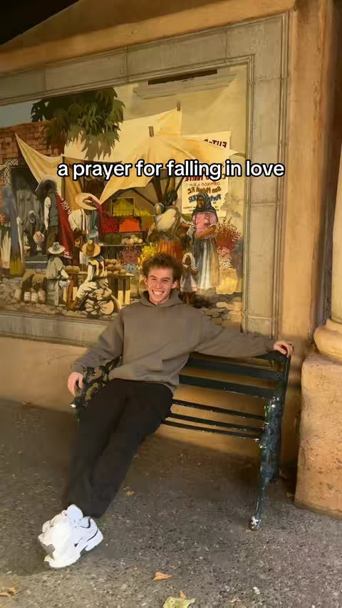 A young man sits on a bench in front of a mural. He's smiling and wearing a gray hoodie, black pants, and white sneakers. The background is a colorful painting with figures and architectural elements.