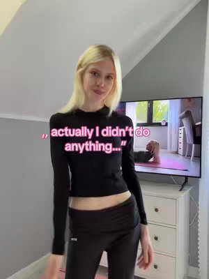 A young woman with long blonde hair, wearing a black top and leggings, stands in front of a TV. The TV screen shows a person doing a pilates exercise. Text overlayed on the image reads, "actually I didn't do anything...". The room has simple decor with a neutral color palette.