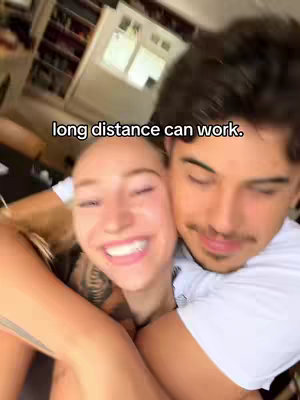 A selfie-style video cover image featuring a smiling woman embracing a man, with a background of a room interior. The woman has blonde hair and visible tattoos on her arm. The man has dark hair and is lightly smiling. Text overlayed on the image reads "long distance can work."