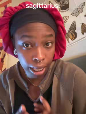A close-up shot of a person's face, who's looking directly towards the camera with a subtle facial expression. The person wears a red head covering, a nose ring, and small hoop earrings. They are wearing a brown hoodie and a black top. Behind them, a wall is decorated with images of butterflies.