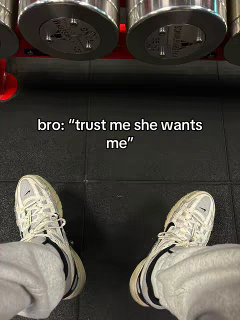 The image shows a first-person view of someone's feet in front of a gym setting. The person is wearing white sneakers with gray accents and black soles. The feet are positioned over a black floor with dark gray tiles. In the background, blurred chrome gym equipment is discernible. Text overlaid on the image reads, 'bro: "trust me she wants me"'.