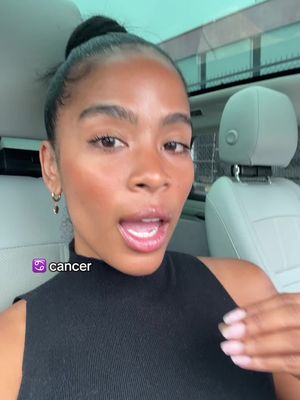 A woman with her hair in a bun is filmed from the chest up while sitting in a car. She has a neutral facial expression with a slightly open mouth, and a small earring is visible. The background looks like the inside of a car. Text overlay in the lower left corner identifies the zodiac sign.