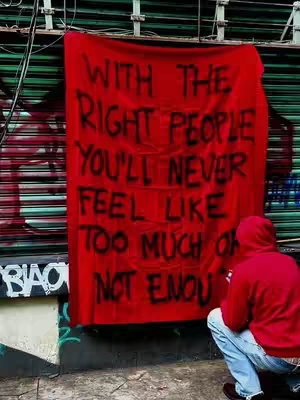 A person in a red hoodie is crouched down in front of a red banner with black text, the banner is hanging in front of a metal shutter. The text on the banner reads: WITH THE RIGHT PEOPLE YOU’LL NEVER FEEL LIKE TOO MUCH OR NOT ENOUGH.