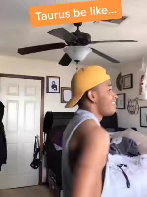 The cover image shows a young man in a yellow hat and gray tank top, looking slightly off-camera with a smile. Above his head is a text overlay in an orange rectangle that says "Taurus be like..." The setting appears to be a room in a home, with a ceiling fan and pictures on the wall visible in the background.