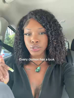 A woman with curly black hair is sitting in a car. She is wearing a black zip-up hoodie and a necklace with a large, green pendant. Text overlayed on the image reads, "Every demon has a look."