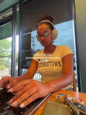 A young woman with curly hair wearing glasses and a beige Houston Rockets t-shirt studies at a table with a laptop. She has headphones on and is typing. The scene takes place in a cafe setting with a window visible in the background.