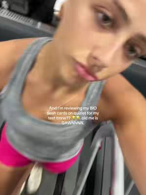 A selfie-style video shot from a slightly low angle in a gym setting. The individual is wearing a grey tank top and pink shorts, and appears to be working out or on a treadmill. The background is blurred, but gym equipment is visible. Below, there's overlaid text from the creator reviewing flashcards for a test.