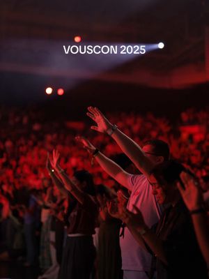 A low angle shot of a large crowd with arms raised in a concert hall or a religious setting, lit with deep red and purple hues from stage lights. On top, a bright white text reads 'VOUSCON 2025'.