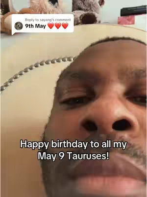 A close-up shot of a Black man centered in the frame, with the top of his head cut off. He has a warm complexion and is looking at the camera. The background includes glimpses of a patterned wall, a sign, and a few stuffed animals. The overlay displays a message: 'Happy birthday to all my May 9 Tauruses!' set against a black bar.