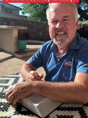 A middle-aged man with gray hair and a beard sits outside, holding a Bible open in front of him. He is smiling and looking toward the camera. The background includes a building, a paved area, and some greenery.