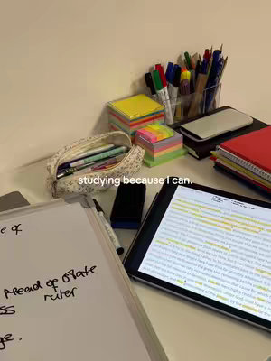 A desk setup with study materials. There's a whiteboard with handwritten notes, a tablet displaying a document with highlighted text, and various stationery items like pens, sticky notes, and notebooks. The color palette is neutral, with a focus on white and beige for the desk and background, and pops of color from the stationery.
