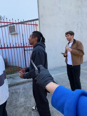 A photograph captures a scene with three individuals in an outdoor setting. A man of color is the central figure, seen from the side, with a concentrated expression and dreadlocks pulled back. He stands facing slightly left, with his arms at his sides. To the right, another man holds a drink and a phone, looking towards the central figure with a neutral expression. The focus is drawn to a black-gloved hand holding what appears to be a pair of tweezers, positioned in front of the face of the central figure. The background features a simple concrete wall and a red fence. A person wearing a blue jacket can be seen from the chest downward, on the far left