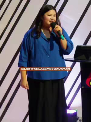 A woman wearing a blue shirt and black skirt stands in front of a stage with a black and white striped background. She is holding a microphone and speaking.